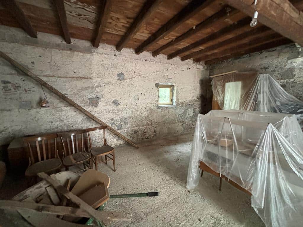 Vecchia soffitta ristrutturata con travi in legno e pareti in pietra, pronta per essere trasformata in uno spazio abitabile, con mobili coperti da teli di plastica per protezione durante i lavori di ristrutturazione.