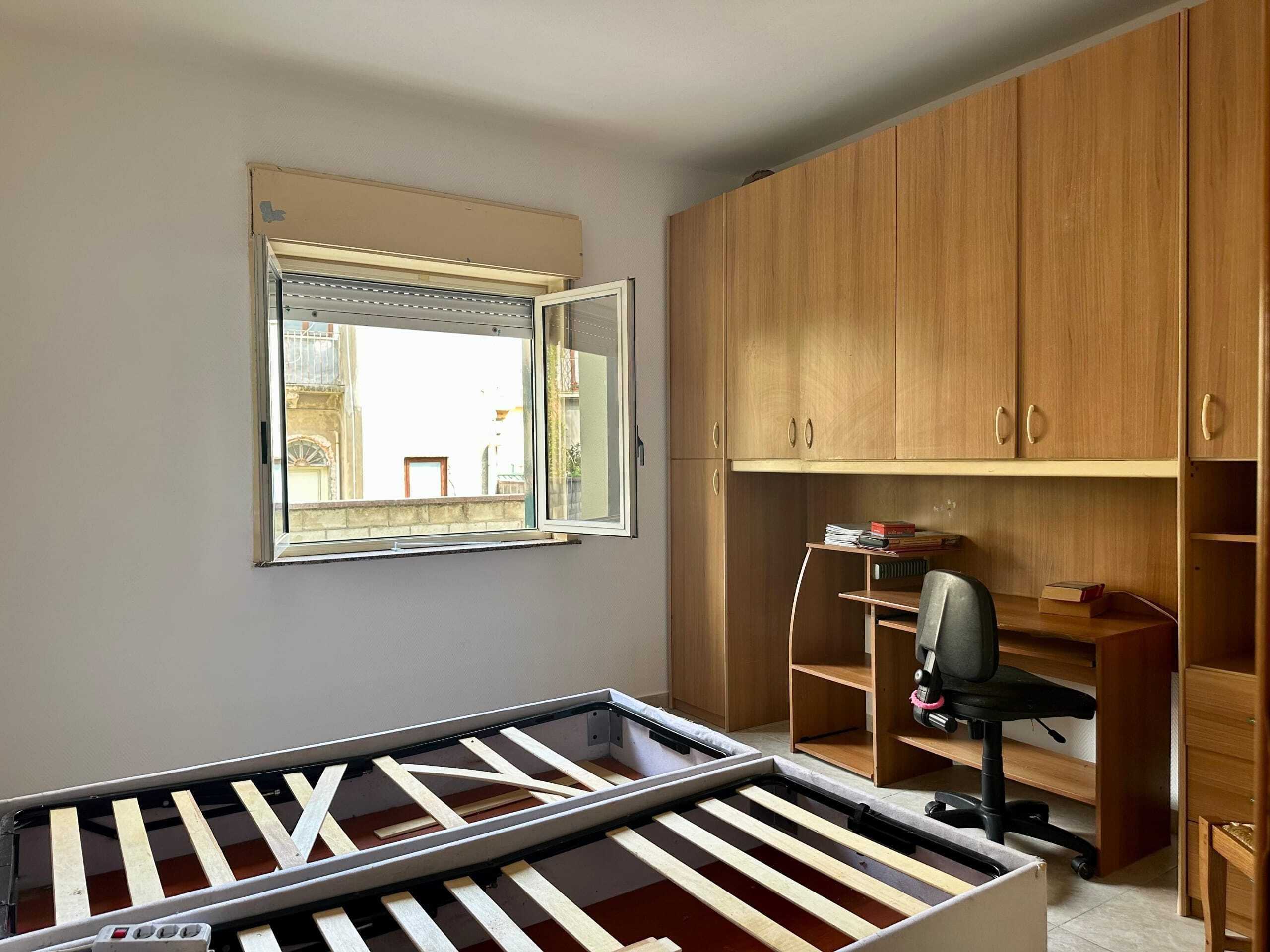 Letto singolo senza materasso e scrivania in una stanza con armadio in legno e finestra aperta, luce naturale che entra nell'ambiente.