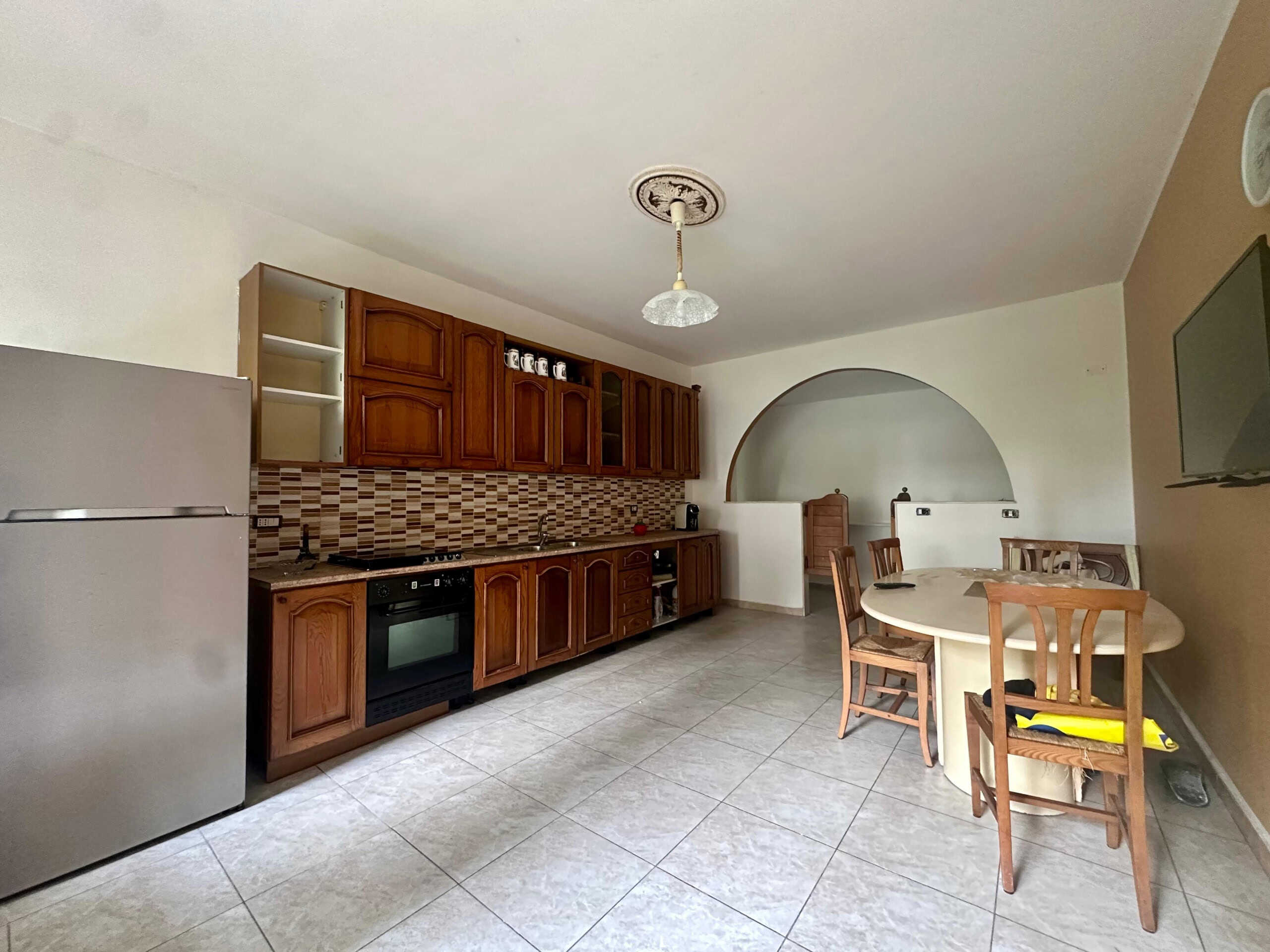 Spazio cucina con mobili in legno, piano cottura e frigorifero in ambiente abitabile, ideale per living o zona pranzo, con tavolo rotondo e sedie in legno.