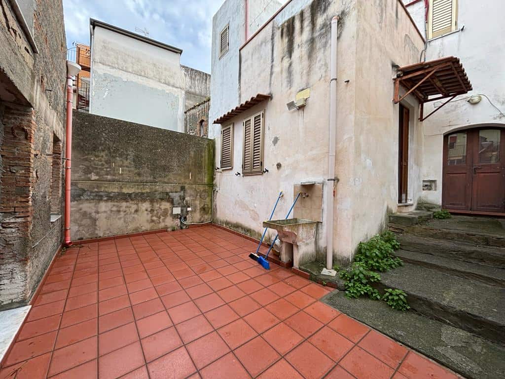 Cortile interno di edificio residenziale con pavimentazione in gres rosso, spazio esterno con accesso alle scale e alle finestre, zona lavanderia con lavabo e scope. Muratura storica e pareti di diverse altezze.