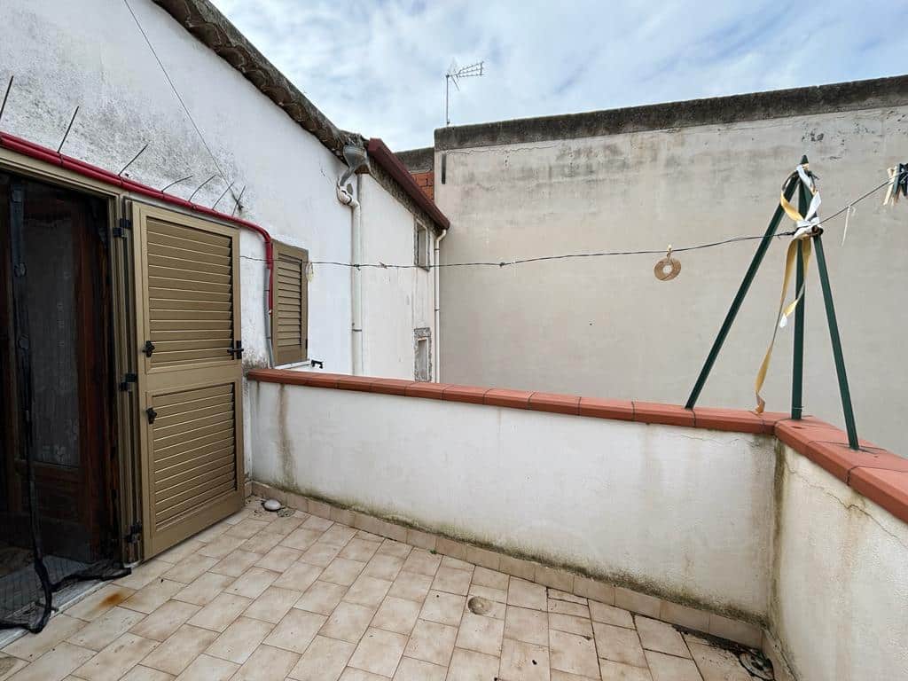 Balcone di un appartamento con muri in pietra e cancelli alle finestre, pavimento in piastrelle beige, corda per stendere e tende a scomparsa, vista sulla parete esterna di un edificio storico, ideale per mangiare o rilassarsi all'aperto.
