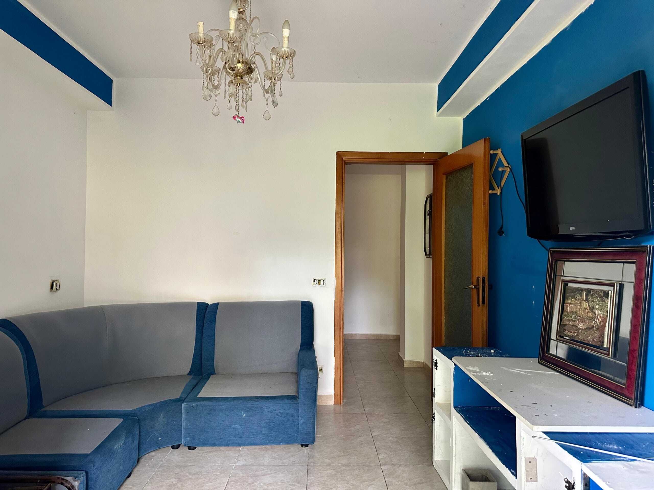 Divano angolare blu in soggiorno con pareti bianche e una parete blu, televisore e quadro decorativo, ambiente tranquillo e funzionale.