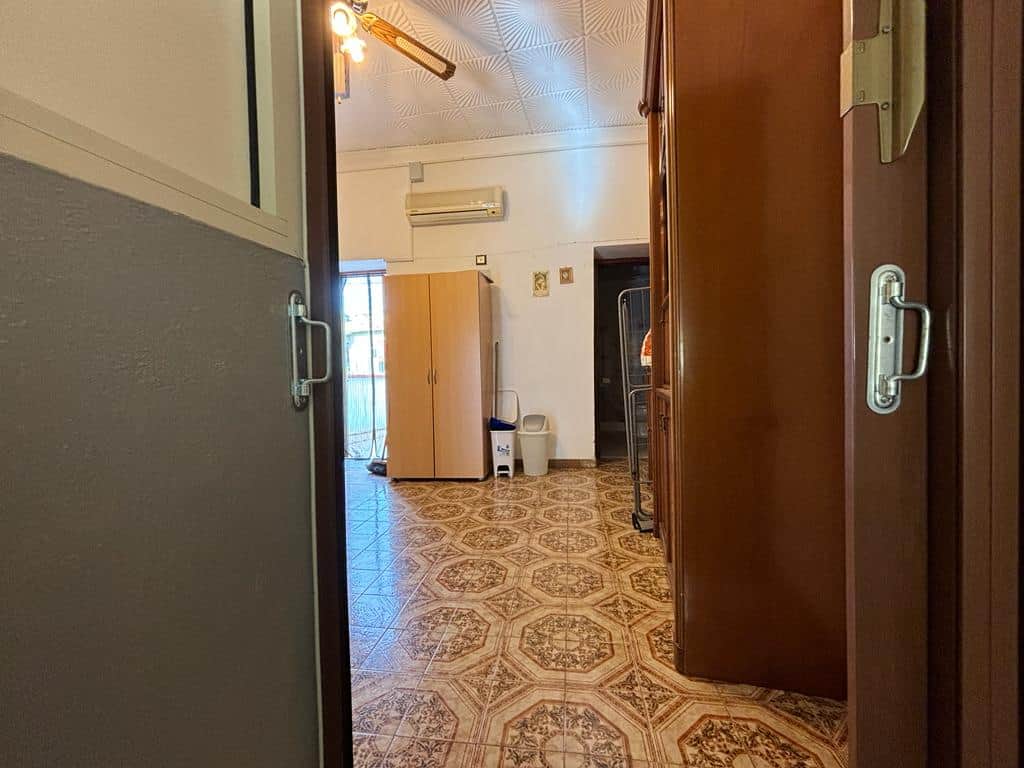 Porta d'ingresso aperta che rivela una stanza con mobili e arredo classici, pavimento in piastrelle decorato, aria condizionata e luce soffusa, stile abitativo tradizionale.