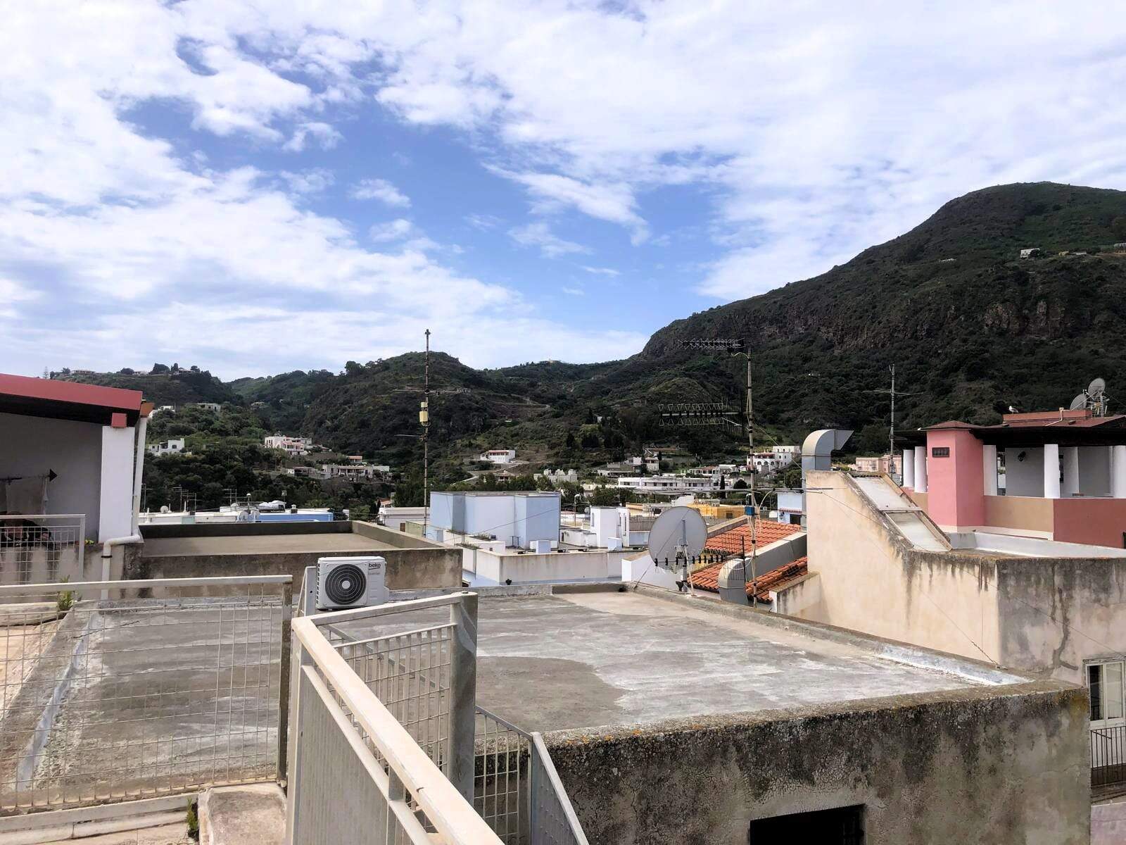 Techo di un edificio con satellite e condizionatori, con vista sulle montagne e case circostanti, cielo sereno con alcune nuvole, ambiente urbano e naturale in Italia.