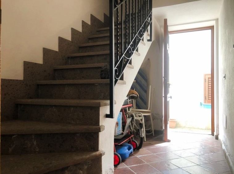 Scalinata interna in pietra con ringhiera in ferro battuto, ingresso di casa con porta aperta, oggetti vari come biciclette e sedie, luce naturale che entra dall’esterno.