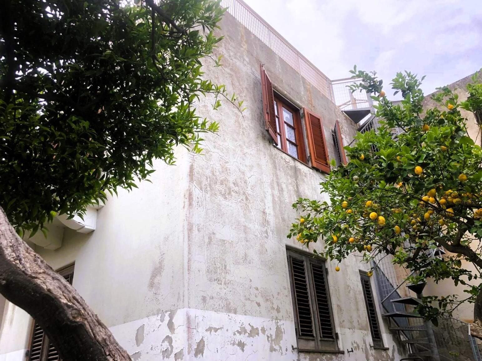 Vecchio edificio con finestra a persiana in legno, pareti in pietra, fontana e limoni in un cortile esterno, fronte strada, atmosfera mediterranea, stile tradizionale italiano.