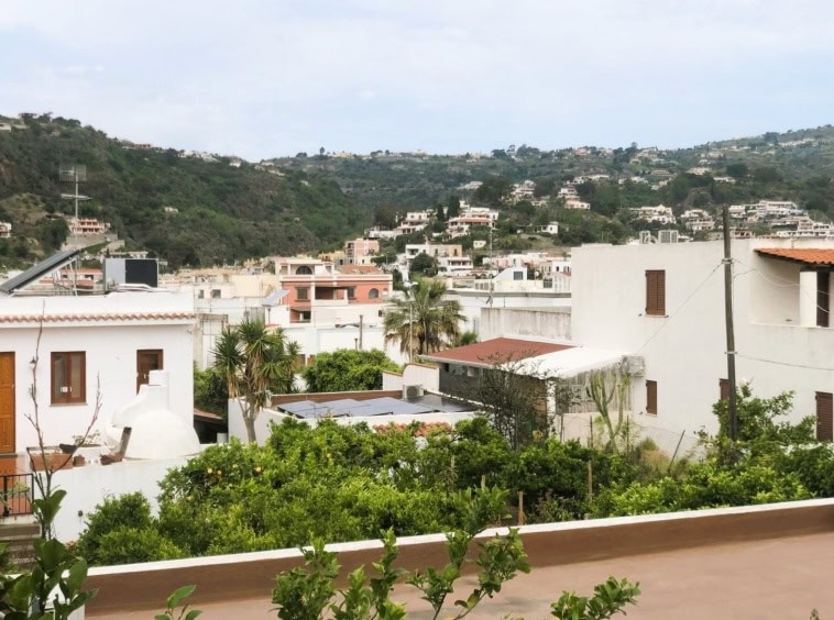 Vista panoramica di case bianche in stile mediterraneo con giardini verdi, su un paesaggio collinare con case e vegetazione, vista sulla costa e sul cielo sereno.