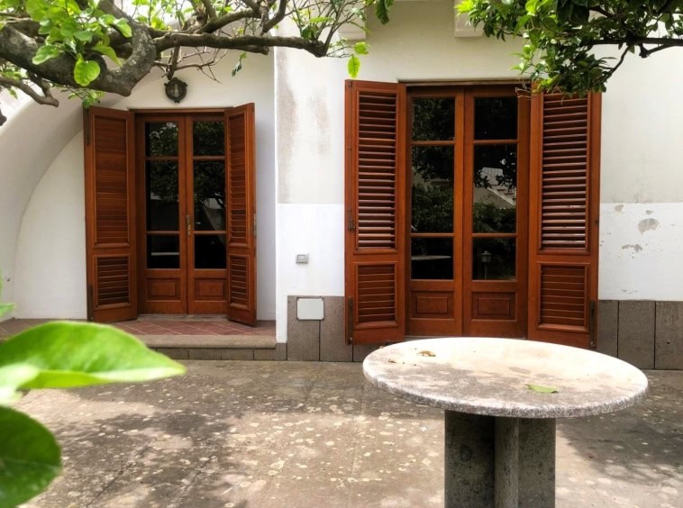 Persiane in legno per porte e finestre con apertura a battente, installate in una casa di campagna, con cortile in pietra e cornice di arbusti verdi, stile rustico e tradizionale.