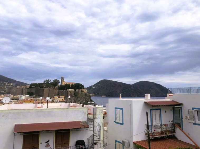 Vista panoramica delle case sul mare con isola e colline sullo sfondo, cielo nuvoloso, suggestivo scenario costiero, ideale per vendita immobiliare a NextCasa, Roma, Italia.