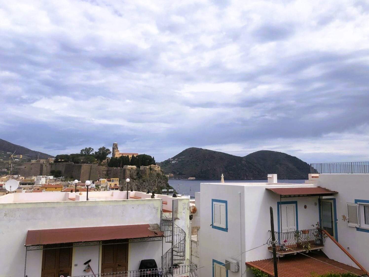 Vista panoramica delle case sul mare con isola e colline sullo sfondo, cielo nuvoloso, suggestivo scenario costiero, ideale per vendita immobiliare a NextCasa, Roma, Italia.