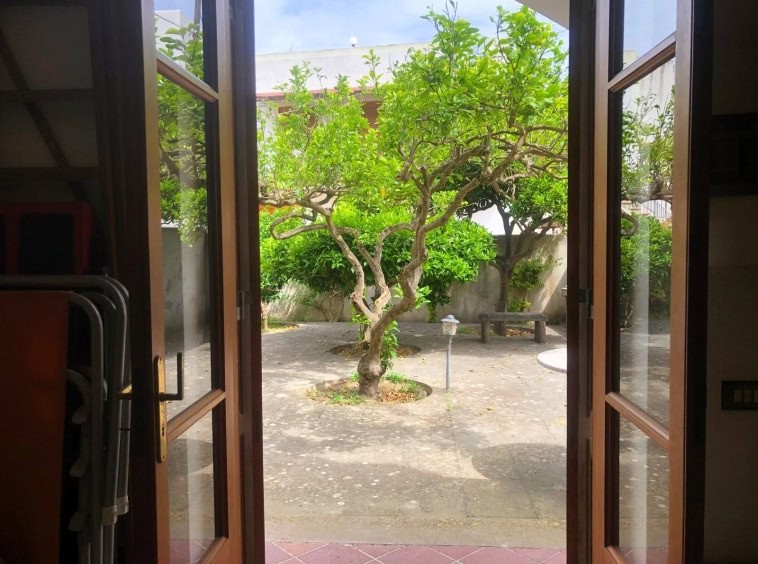 Finestra aperta che rivela un cortile interno con alberi verdi e panchine, ideale per vivere momenti di relax all'aperto in contesti residenziali o case con giardino, perfetto per immobili in vendita o affitto.