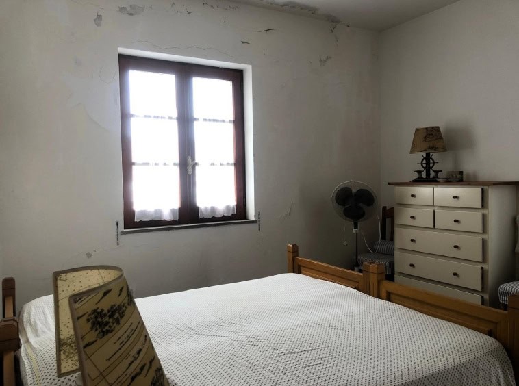 Vecchio stile camera da letto con finestra di legno, pareti con segni di rustico, arredamento semplice e funzionale, fra cui comò, lampada e ventilatore, in un interno da ristrutturare o restaurare.