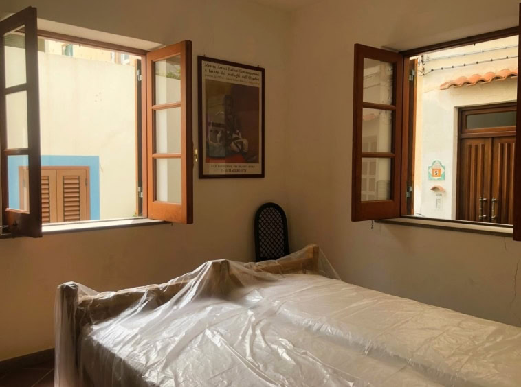Letto con copriletto di plastica in una stanza luminosa con doppie finestre in legno, vista aperta sulle facciate di case diverse in un ambiente domestico.