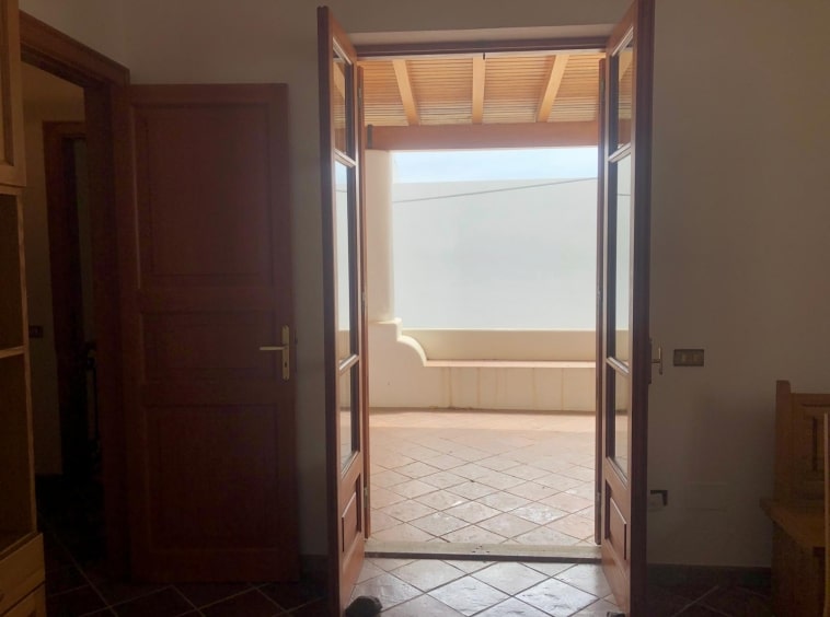 Porta d'ingresso in legno che si apre su un balcone esterno con vetrate, illuminato dalla luce naturale in un ambiente residenziale.