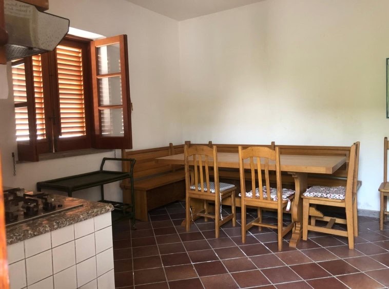 Ampia sala da pranzo con mobili in legno, cucina con stufa a gas, finestre con persiane di legno, pavimento in piastrelle marroni, ambiente luminoso e accogliente in stile rustico.
