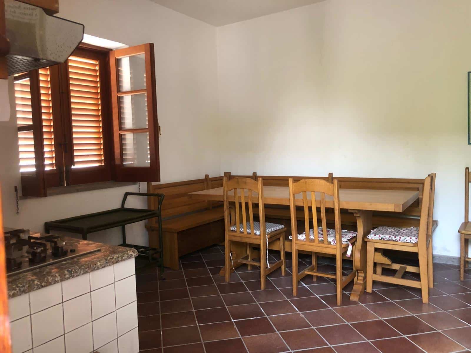 Ampia sala da pranzo con mobili in legno, cucina con stufa a gas, finestre con persiane di legno, pavimento in piastrelle marroni, ambiente luminoso e accogliente in stile rustico.