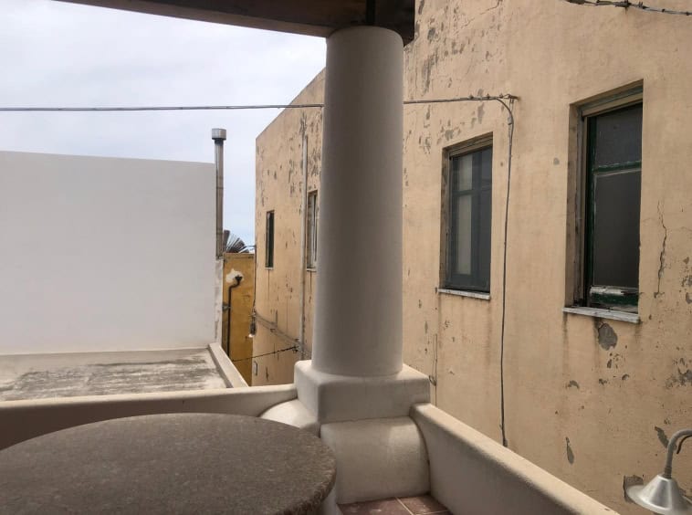 Balcone con vista su edificio antico con pareti usurate, finestra con persiana verde, colonna bianca, cielo nuvoloso, elementi di arredo in pietra e tubi esterni.