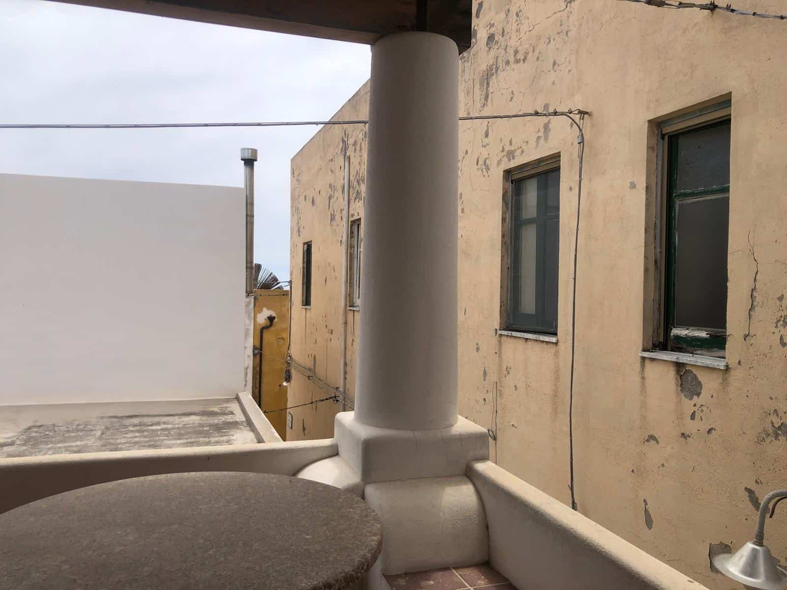 Balcone con vista su edificio antico con pareti usurate, finestra con persiana verde, colonna bianca, cielo nuvoloso, elementi di arredo in pietra e tubi esterni.