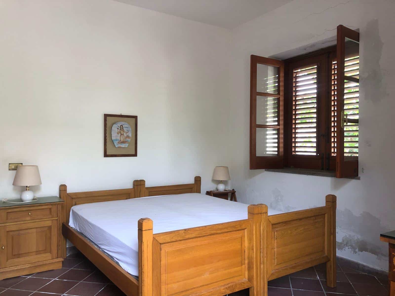 Letto matrimoniale in camera con pareti bianche e finestra con ante di legno, arredamento rustico, stile country, illuminazione naturale, atmosfera accogliente, case vacanza, affitti turistici, NextCasa.