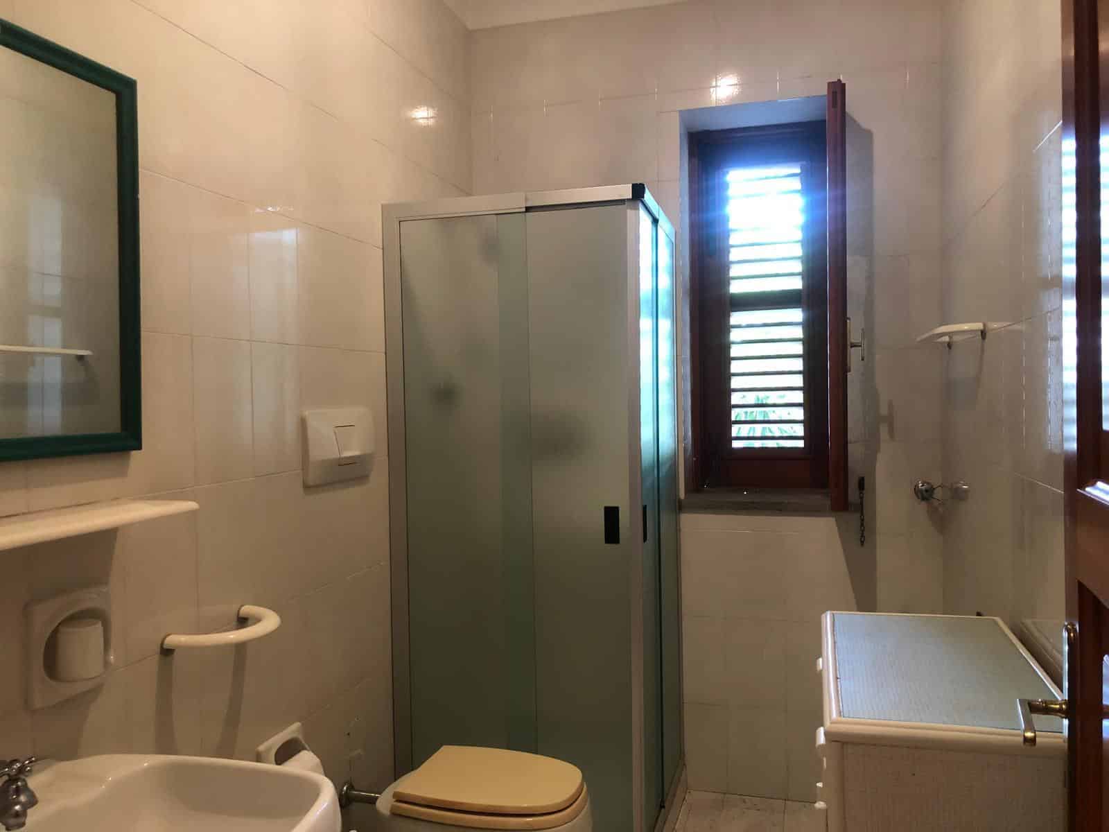 Bagno con finestra, sanitari, piastrelle chiare, doccia, armadietto, arredamento semplice, luce naturale, stile classico, bagno funzionale, ambiente luminoso.