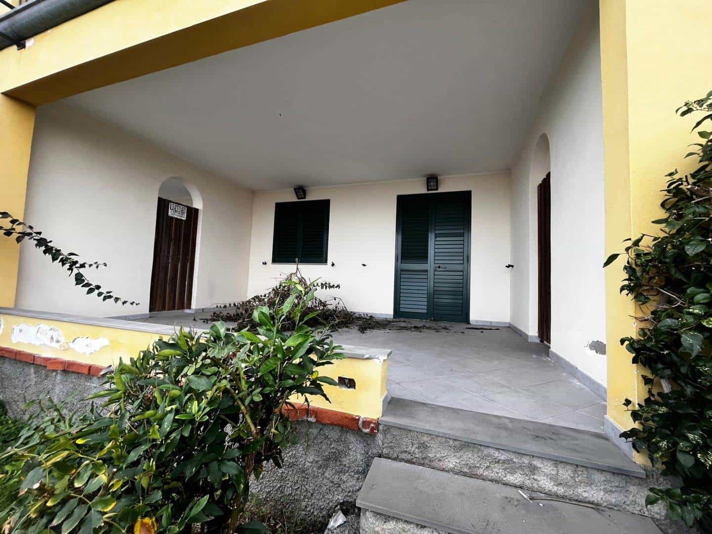 Portico di casa con porta e finestre in legno verniciato, ambiente esterno con piante verdi e recinzione in pietra, casa in stile tradizionale, spazio accogliente e ben curato.