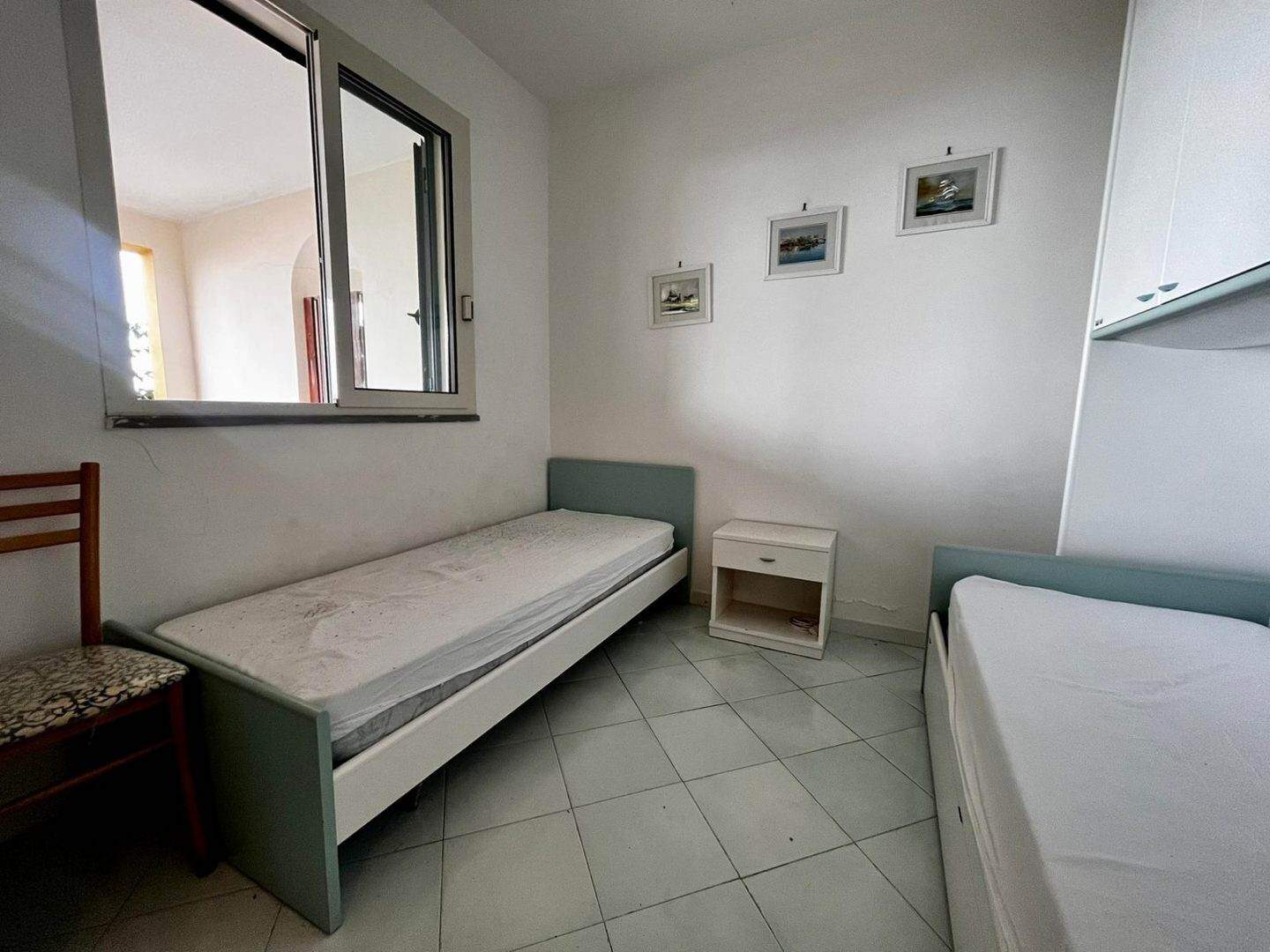 Stanza singola in affitto con letto singolo, arredi essenziali, pareti bianche e finestra con vista su balcone, ambiente luminoso e semplice, ideale per studenti o lavoratori in cerca di sistemazione temporanea.