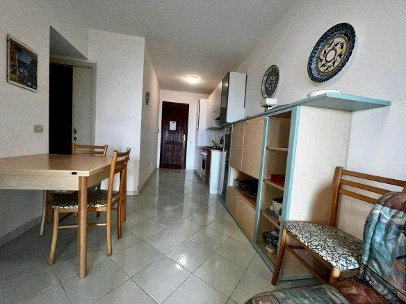 Spazio soggiorno con cucina e area pranzo, arredamento semplice, pareti bianche, piastrelle lucide, mobili in legno e moduli di stoccaggio colorati, ambiente luminoso e accogliente.