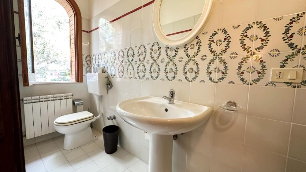 Lavabo e servizi igienici in bagno con mosaico decorativo, finestra e riscaldamento a radiatori, ideale per servizi di immobili residenziali e commerciali.