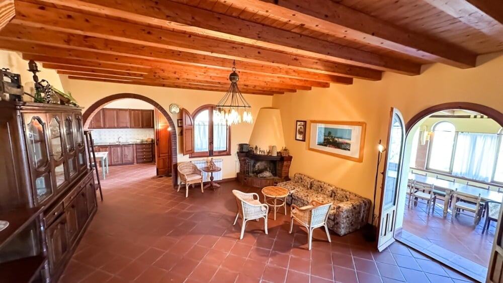 Elegante soggiorno rustico con soffitto a travi in legno, divano, camino e zona pranzo vicino al balcone, perfetto per relax e accoglienza in stile tradizionale toscano.
