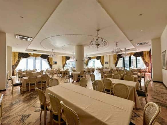 Ampia sala ristorante con arredi eleganti, grandi finestre, tende drappeggiate e dettagli decorativi in stile classico, ideale per eventi e cene in ambienti raffinati.