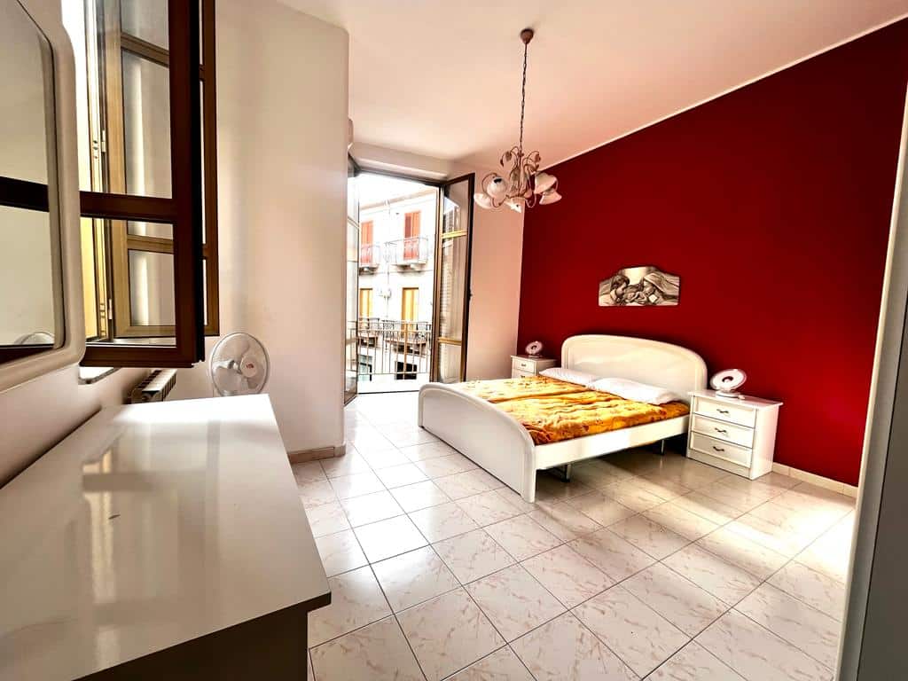 Ampia camera da letto luminosa con letto matrimoniale, mobili moderni, parete decorativa rossa, balcone con vista su edificio storico, arredamento elegante, ideale per affitto o vendita immobiliare NextCasa.