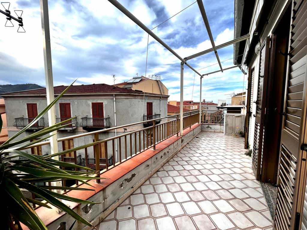 Balcone con vista su tetti e cielo nuvoloso, pavimentato in piastrelle beige e bordo in terracotta, dotato di struttura per stendere e piante verdi, ideale per spazi esterni di case o appartamenti NextCasa.