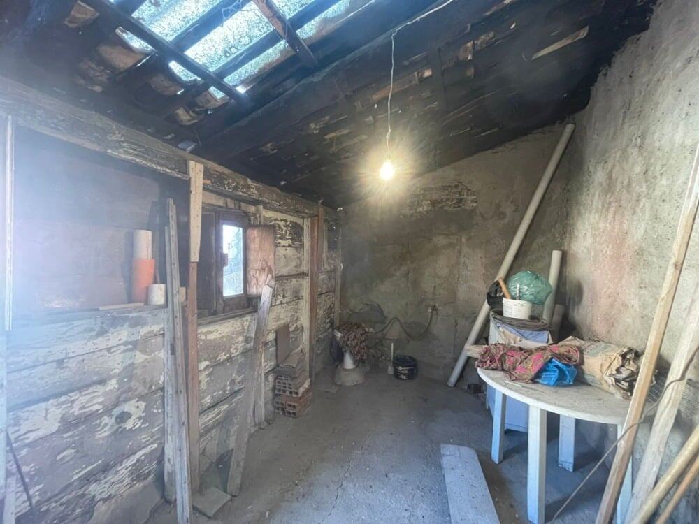 Altezza soffitto con travi di legno e muro in pietra grezza, interno di una stanza rustica in ristrutturazione, con attrezzi da lavoro e materiali edili, illuminata da una singola lampadina.