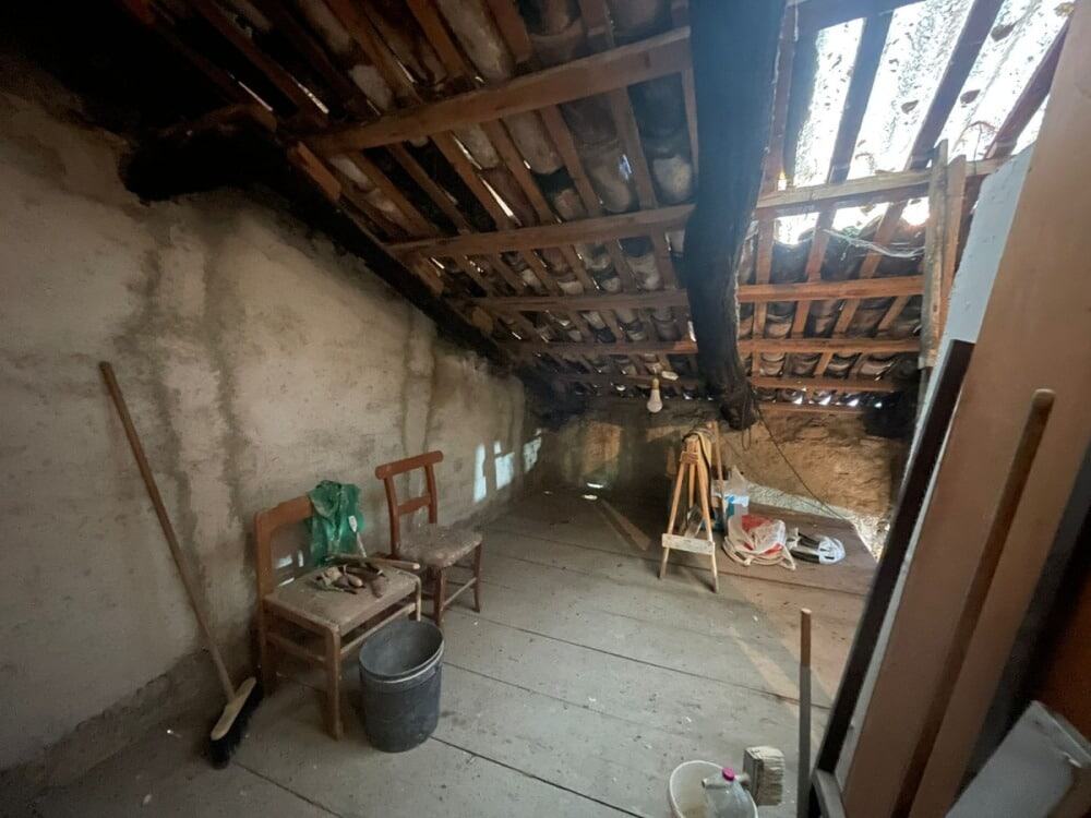 Vecchio sottotetto con tetto in legno, pareti in pietra e pavimento in legno, contenente sedie, secchio, attrezzi da lavoro e materiale di costruzione, in fase di ristrutturazione o conservazione rustica.