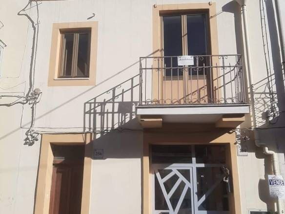 Finestra e balcone di una casa residenziale in stile mediterraneo, facciata bianca con dettagli beige, porta d'ingresso moderna e un cartello "Vende" visibile, in un contesto urbano soleggiato.