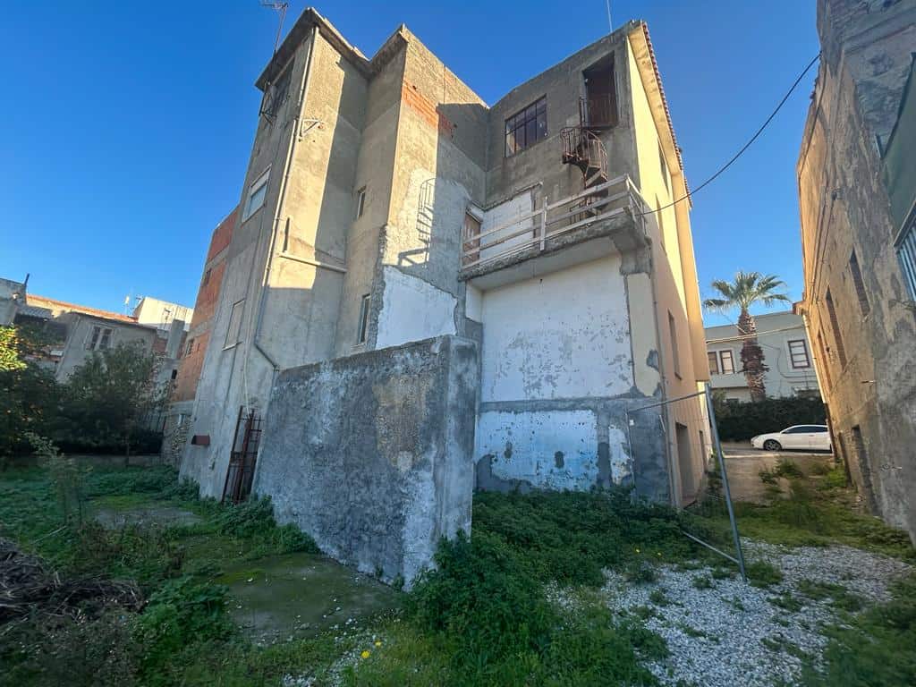 Vecchio edificio da ristrutturare con pareti in cemento e balcone in ferro battuto, situato in zona urbana con spazio verde e strada asfaltata nei pressi.