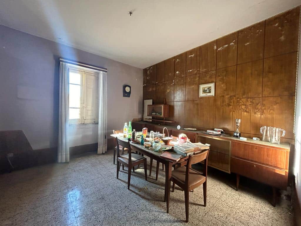 Vecchia sala da pranzo con tavolo apparecchiato e mobili in legno, pareti rivestite di pannelli di legno e finestra con tende bianche, ambiente di casa da ristrutturare.