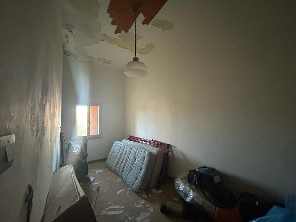 Immagine di una stanza in fase di ristrutturazione con pareti bianche, soffitto danneggiato, finestra che lascia entrare luce naturale, mobili e vecchi oggetti accatastati, ambiente da rinnovare.