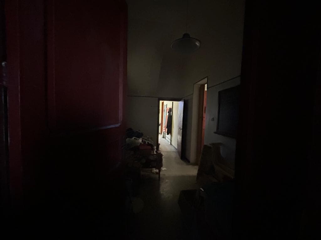 Immagine di un interno abitazione con poca illuminazione, porta di ingresso sullo sfondo e arredi vecchi e disordinati, rappresentativa di immobili da ristrutturare o in condizioni di bisogno di rinnovamento.