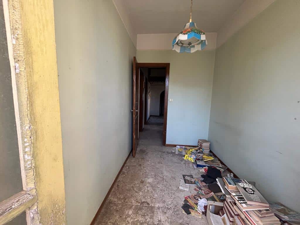 Rilievo di una stanza da ristrutturare con pareti verdi e pavimento in cemento, disordine con libri e riviste, porta in legno e lampada a soffitto colorata, ambiente da sistemare e arredare.