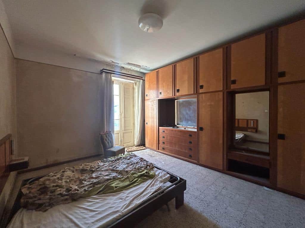 Vecchio soggiorno con armadio in legno, letto disordinato e finestre con tende, ambiente abitativo da ristrutturare per rinnovare gli interni.