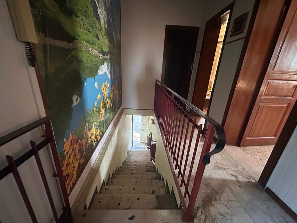 Scalinata interna di una casa con parete decorata da un grande quadro paesaggistico, ringhiera in legno e vista sulla zona giorno al piano inferiore, ambientazione elegante e luminosa.