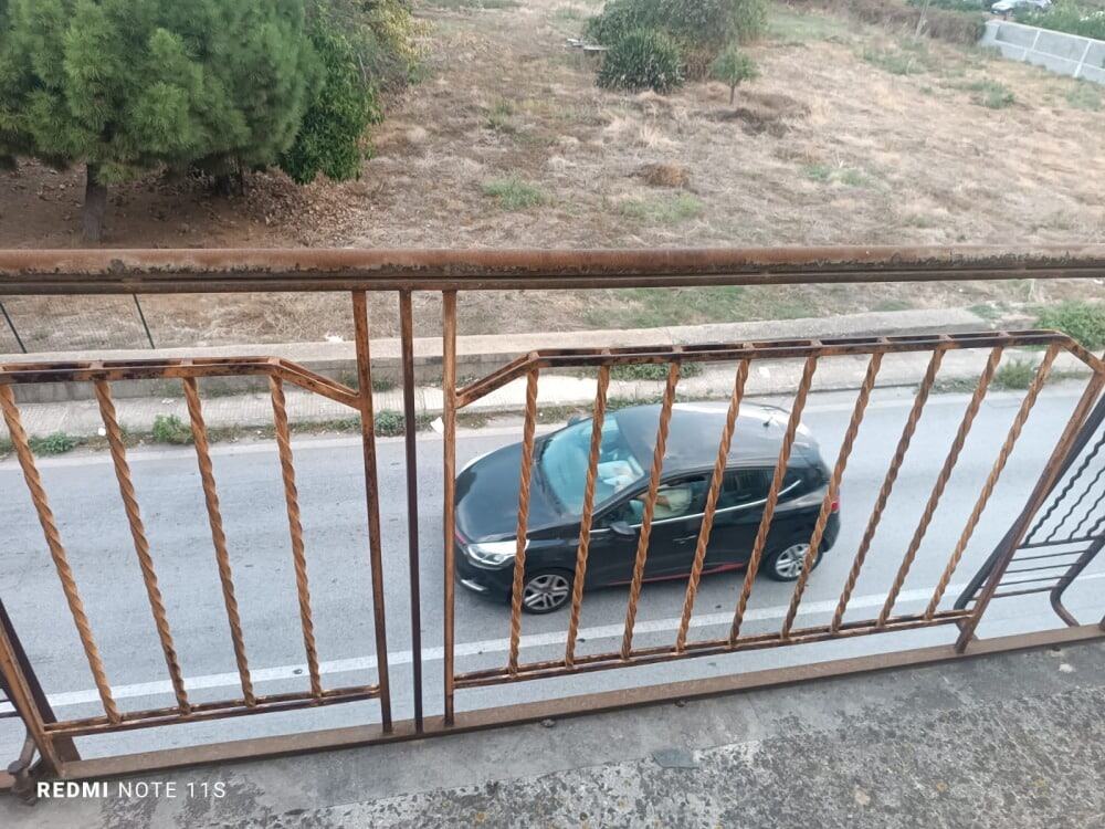 Cancello in ferro battuto con ringhiera stilizzata, vista strada con auto nera e ambiente naturale sullo sfondo, ideale per sicurezza e arredo cortile.