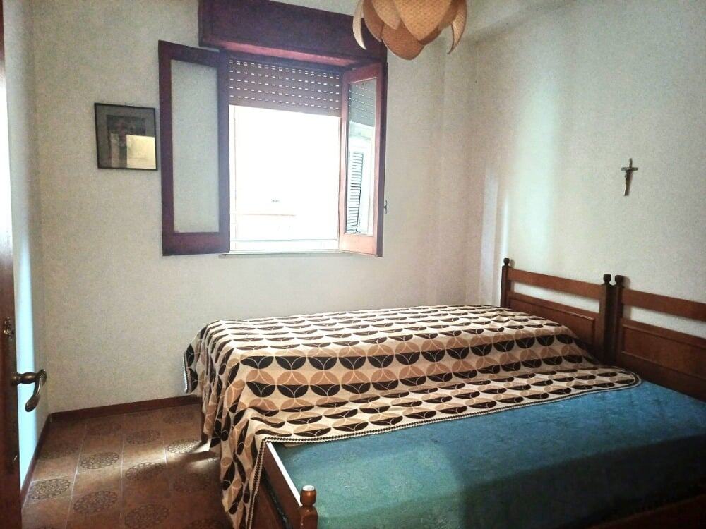 Letto matrimoniale in camera da letto con parete bianca e finestra con tende in legno, decorata con croce e quadro. Stile semplice e funzionale, ideale per ambienti residenziali in stile classico o rustico.