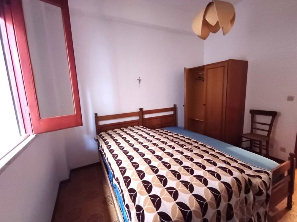 Letto matrimoniale con lenzuola geometriche in una camera semplice e luminosa, con armadio in legno, finestra con persiana rossa e lampada sospesa, ideale per case in affitto o vendita.