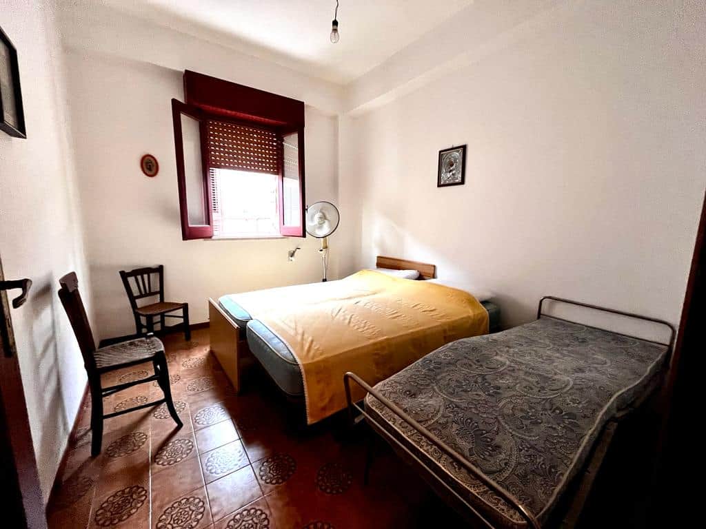 An image of a simple bedroom with two beds, one double and one single, featuring basic furniture, a window with red shutters, and minimal decor, ideal for affordable accommodation.