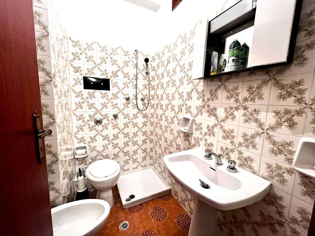 Lavabo e accessori bagno in stile vintage in bagno piccolo con pareti rivestite di piastrelle floreali, dotato di doccia, WC e bidet, arredo funzionale, soluzione ideale per spazi limitati.