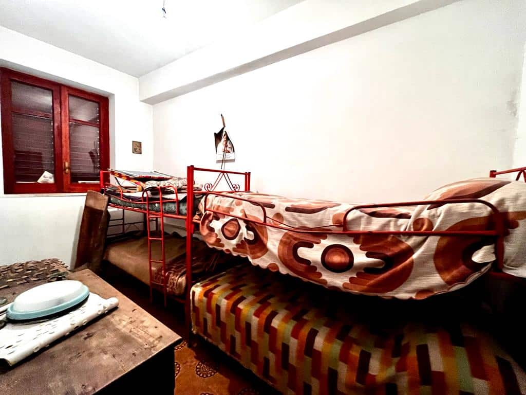Letto a castello in una stanza con pareti bianche e finestra con anta in legno rossa, letto con rete rossa, lenzuola con motivo geometrico, arredamento semplice e atmosfera rustica.