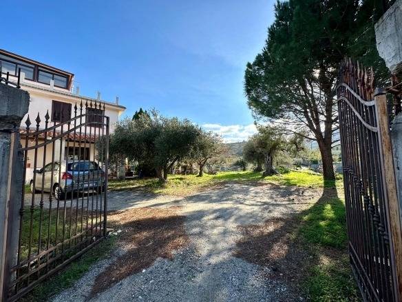 Esito, panoramica di un giardino privato con alberi e spazio verde, ingresso con cancello in ferro battuto, casa residenziale moderna in contesto rurale, atmosfera tranquilla e naturale in Italia.