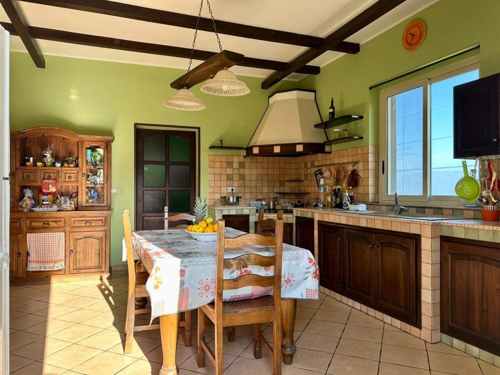 Rustico in stile country con cucina abitabile, pareti verdi, mobili in legno massello, zona pranzo con tovaglia floreale, ampia finestra naturale, ambiente accogliente e ben illuminato.